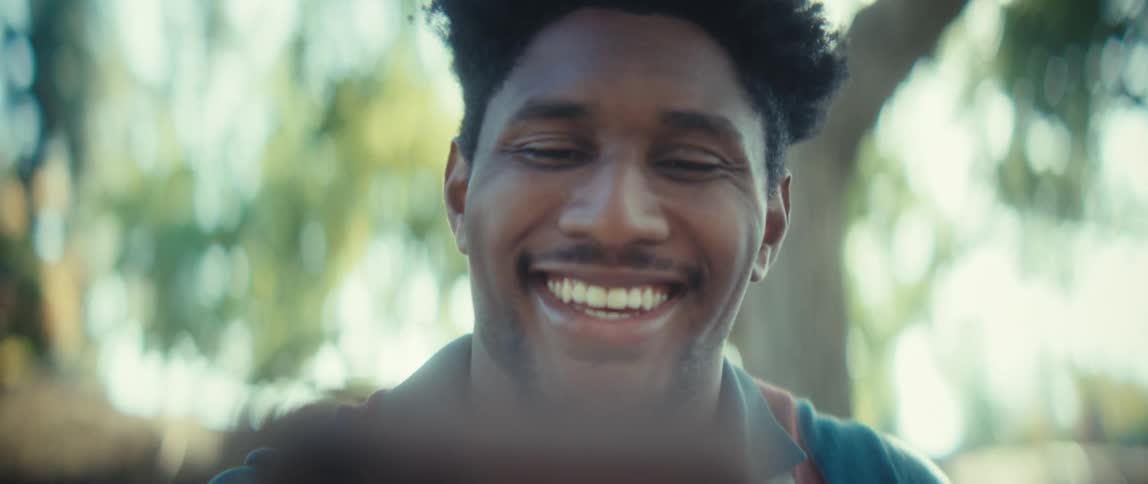 A close view shows a young black man with short dark hair looking down then smiling broadly and waving at a smartphone in bright outdoor sunlight with a blurred green tree canopy in the background