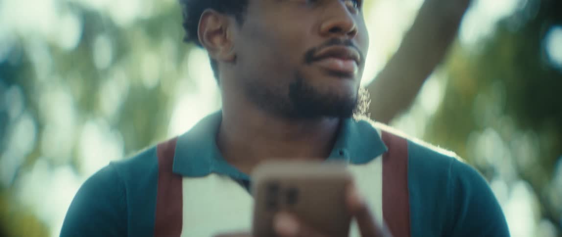 A closeup view shows a man wearing a teal polo shirt and suspenders looking to the side as he holds and uses a smartphone in bright sunlit outdoor surroundings