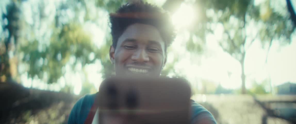 A young black man with dark curly hair smiles and waves while holding a smartphone to record himself for a video chat or selfie with bright sunlight and green trees in the background
