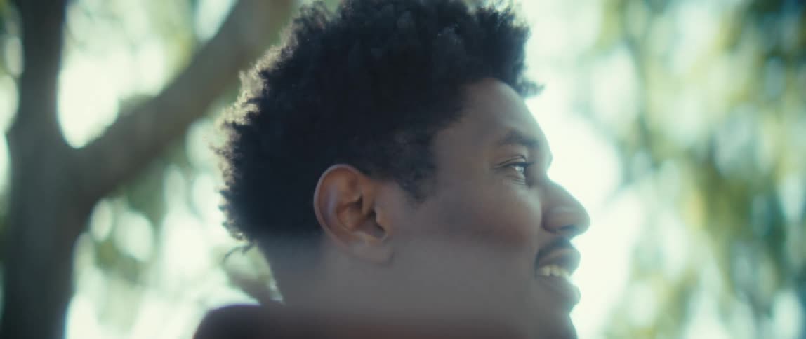 A close view shows a young black man with short curly hair looking talking on his smartphone in the bright daylight with light green blurred foliage in the background