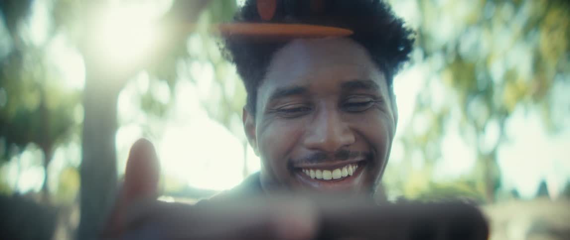 A close view shows a young black man with short dark hair looking into a smartphone held out of frame then smiling brightly with sun flare and green leaves visible in the background