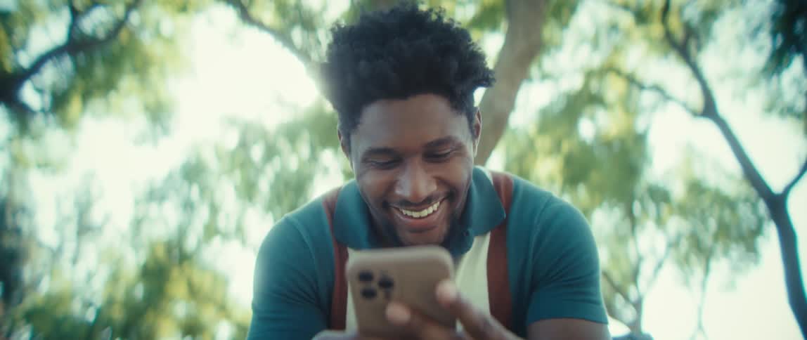 A young man sitting on a bench and laughing while texting on a smartphone with trees behind him