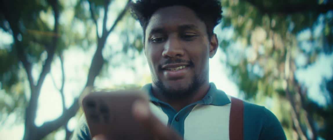 A young man with dark curly hair wears a teal polo shirt with brown suspenders and looks down smiling as he scrolls on a smartphone outdoors beneath the green leaves of trees