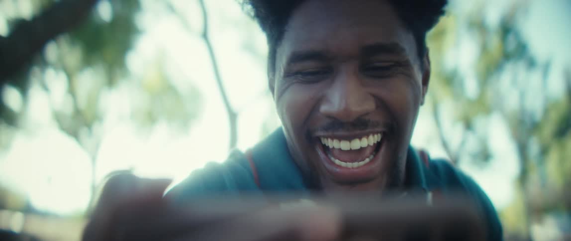 A young man holding a smartphone and laughing and raising hands against a bright outoffocus background of sunlit trees