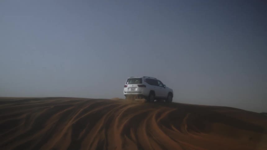 A white suv drives up a large sweeping sand dune under a hazy blue sky leaving tire tracks in the orangebrown desert landscape