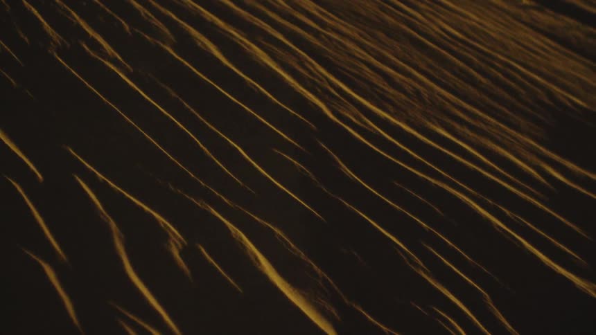A close view tracks over dark desert sand dunes illuminated by warm light catching the sharp linear ridges and creating a striking pattern of light and shadow
