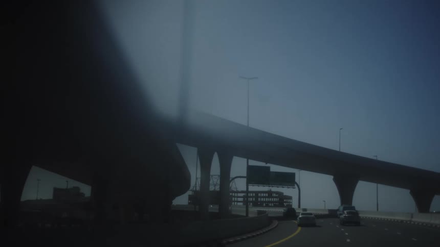 A moving vehicle driving on a wide highway that is passing underneath a massive curving elevated structure supported by thick columns