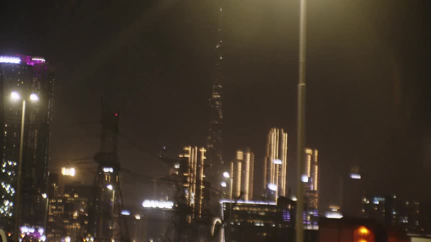 A brightly illuminated vertical pole and several vertically striped towers with many other city lights visible at night against a dark sky