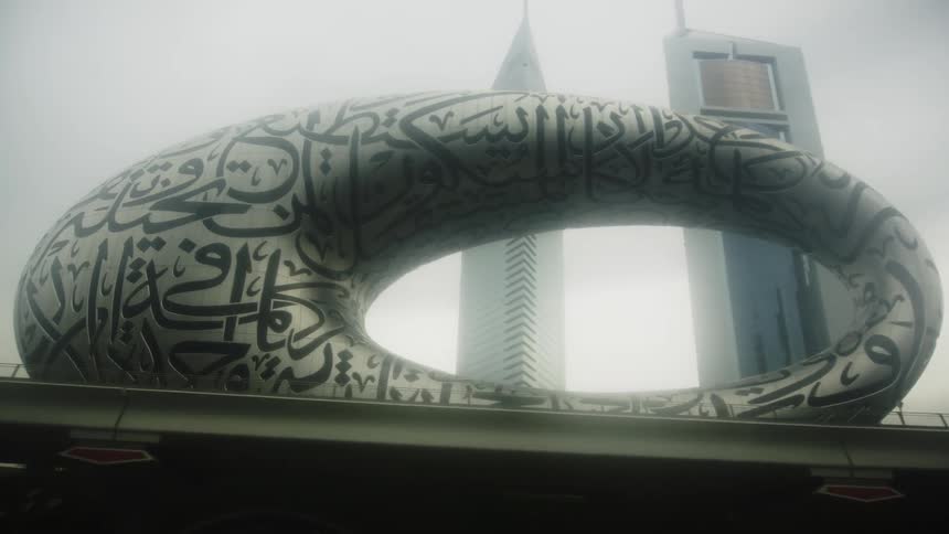 A lowangle view shows the museum of the future in dubai a silver torusshaped building covered in arabic calligraphy standing above an overpass with other skyscrapers visible in the misty background