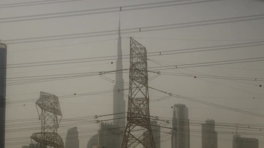 A landscape where massive metal power transmission towers and numerous overhead wires dominate the foreground with several tall structures