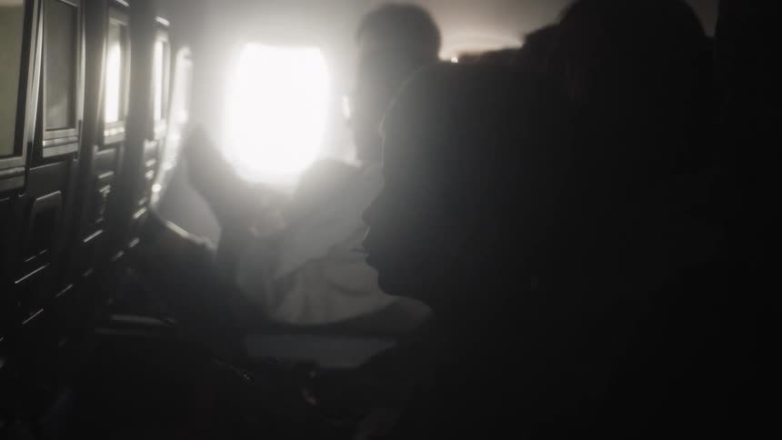 Inside a dim airplane cabin silhouetted passengers sit quietly while bright window light washes over the seats