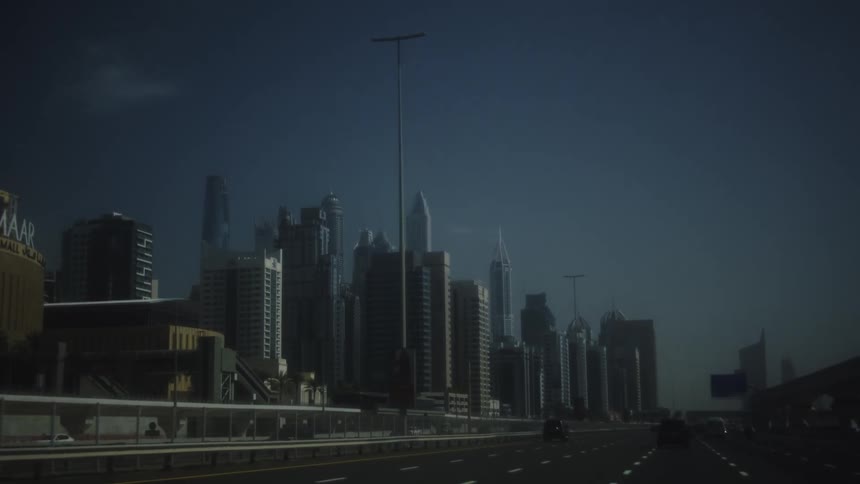 A row of tall buildings in an urban area is viewed from a moving vehicle traveling on a multilane highway under a dark sky
