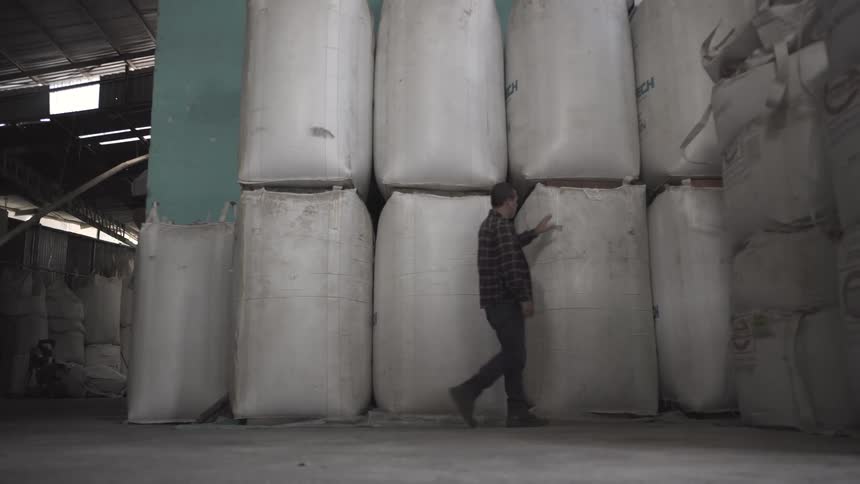 A man wearing a plaid shirt walks past tall stacks of large white industrial sacks in a dimly lit warehouse with bright light visible through the doorway