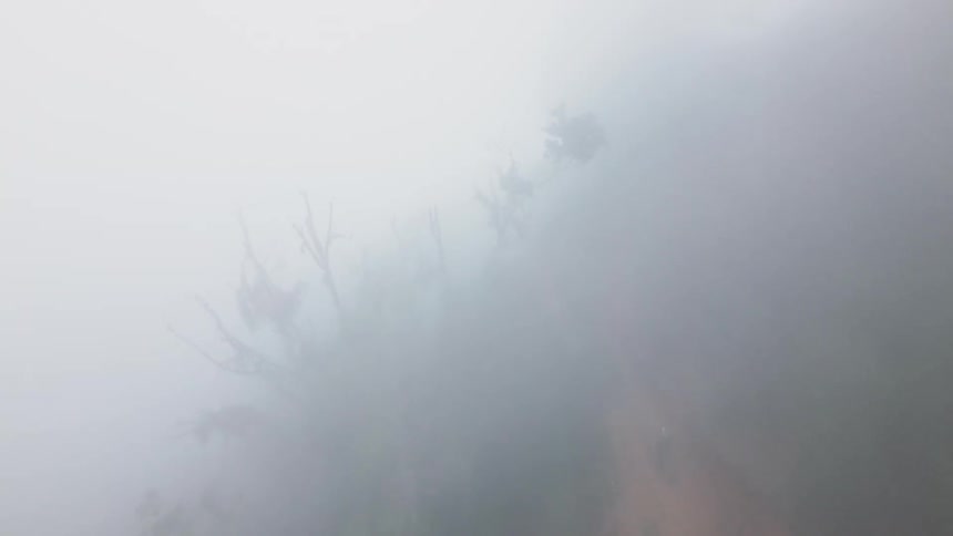 An aerial view shows a biker wearing black and yellow clothing riding an offroad motorcycle on a steep brown dirt trail that runs through a dense green jungle shrouded in thick white fog