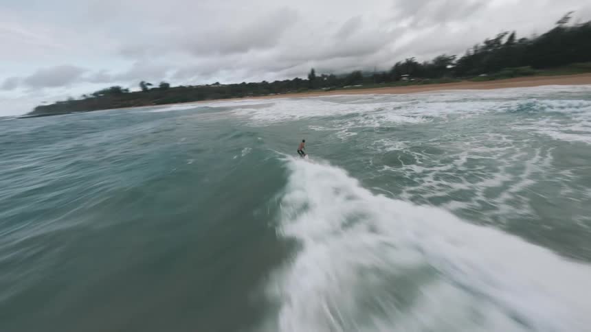 An action aerial view follows a man wearing shorts as he surfs a large turquoise ocean wave that breaks toward a sandy tropical beach under a cloudy overcast sky