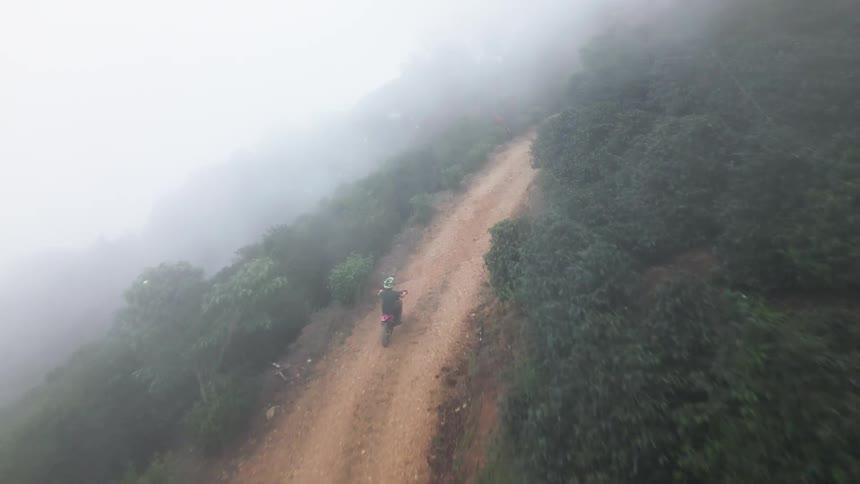 An aerial shot looks down on a person wearing bright green and black gear riding a motorcycle up a steep winding dirt road lined with dark green foliage and obscured by heavy mist