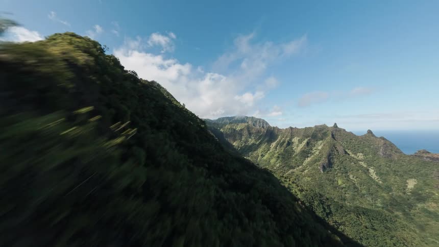 A fast aerial shot sweeps past steep razorsharp mountains covered in dense green jungle foliage revealing more mountain peaks and the blue ocean in the distance under a bright blue sky with white clouds