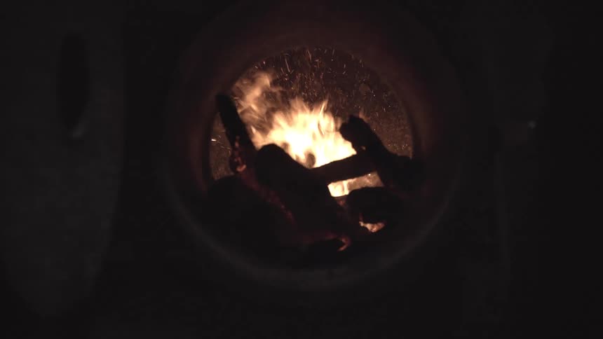 Orange flames roar inside a circular furnace door sparks curling around charred logs in the dark