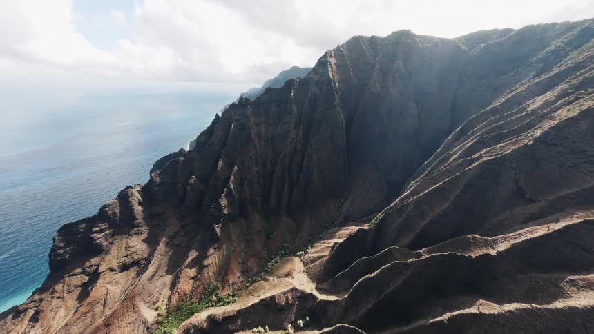 An aerial shot sweeps along the vast reddishbrown cliffs of a steep mountainous coastline revealing the deep blue ocean below and a small boat cutting through the water under a bright sky with scattered white clouds