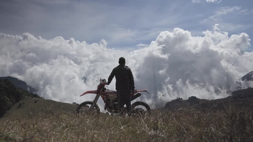 A rider stands beside a dirt bike on a grassy ridge staring out over a sea of billowing clouds under a bright blue sky