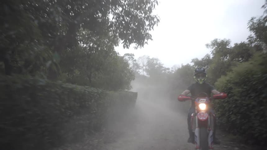 A motorcyclist wearing a helmet and goggles rides a red dirt bike down a muddy foggy dirt road lined with dense green bushes kicking up spray as they move toward the camera