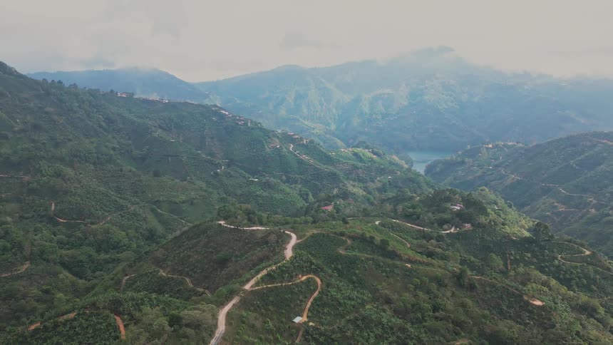 A slow backwardmoving aerial shot captures a vast green mountain range with winding roads and buildings dotting the hillsides