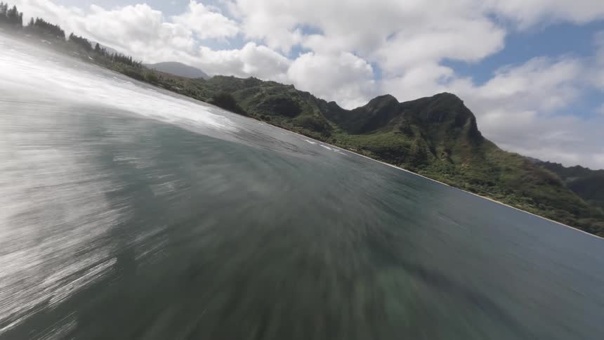 Aerial view of ocean flowing toward steep green mountains under clouds