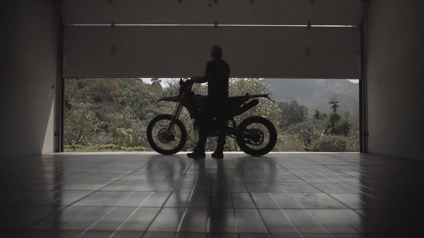 A garage door slowly opens revealing the silhouette of a man wearing a baseball cap standing with a dirt bike inside a dark garage and facing a brightly lit mountainous green landscape