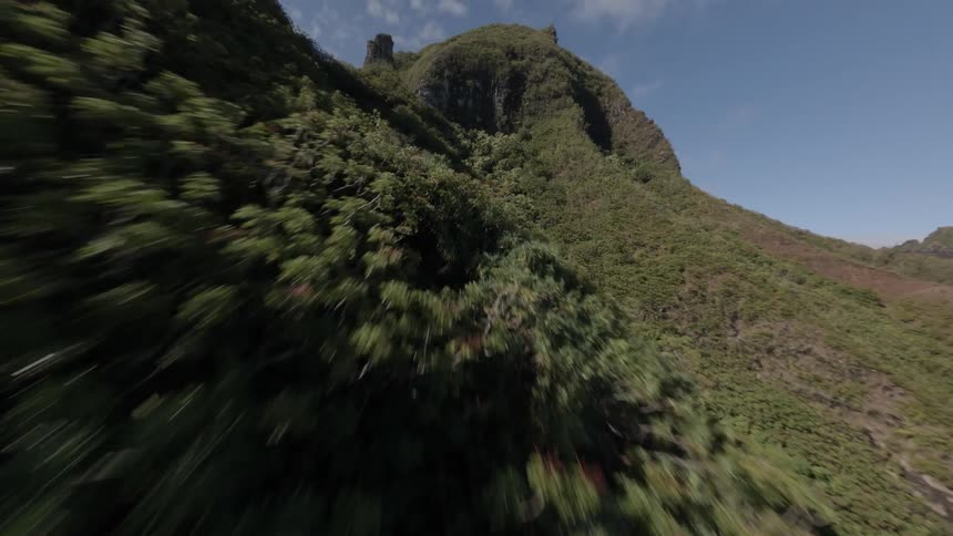 A fastmoving aerial shot descends rapidly past a steep tall mountain face covered in dense green jungle vegetation with a rugged peak visible against a clear blue sky