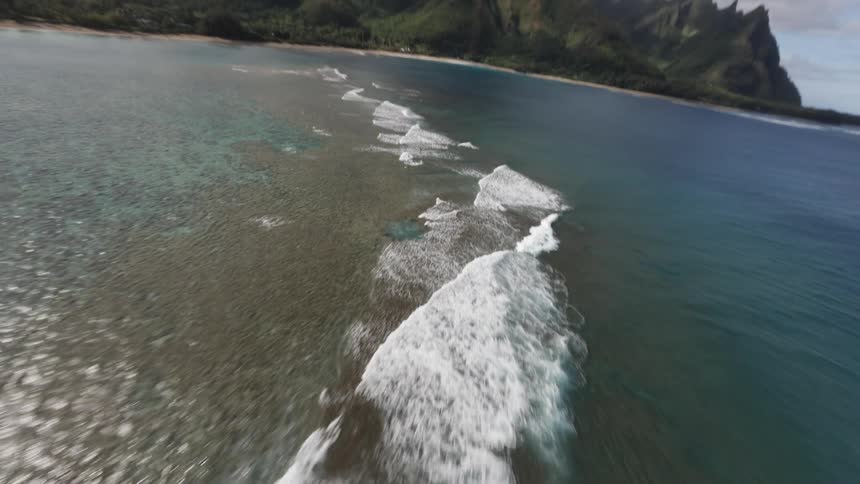 An aerial camera drops down fast from a topdown view to fly at high speed just over the ocean waves towards a mountainous coastline
