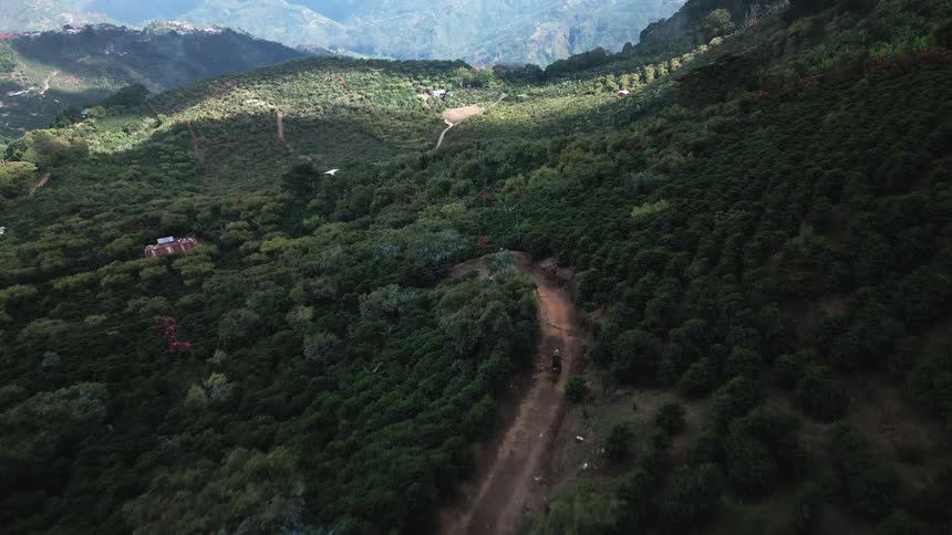Aerial view slowly moving over a densely vegetated mountainous landscape with a dirt road traversing the slopes showing a motorized vehicle moving downward along the path