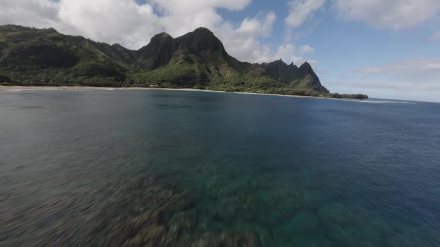 Highangle aerial view rapidly moving backward from a shallow reef with breaking waves toward the open water revealing a wide bay flanked by steep forested mountains under a cloudy blue sky