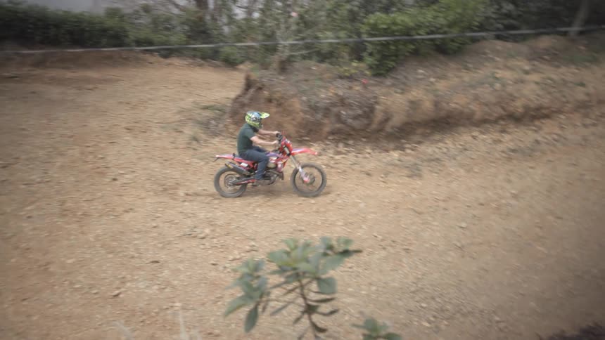 A dirt biker drives into a turn on a dusty track carved into a hillside with a parked car and some scrubby trees nearby under a hazy sky