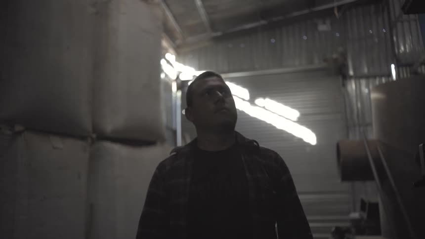 A man wearing a plaid shirt and a dark tshirt walks in a dimly lit industrial warehouse looking up and around with tall stacks of white sacks and corrugated metal walls in the background