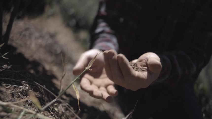 A person kneels on the ground picks up dirt in a hand then throws the dirt down and wipes hands