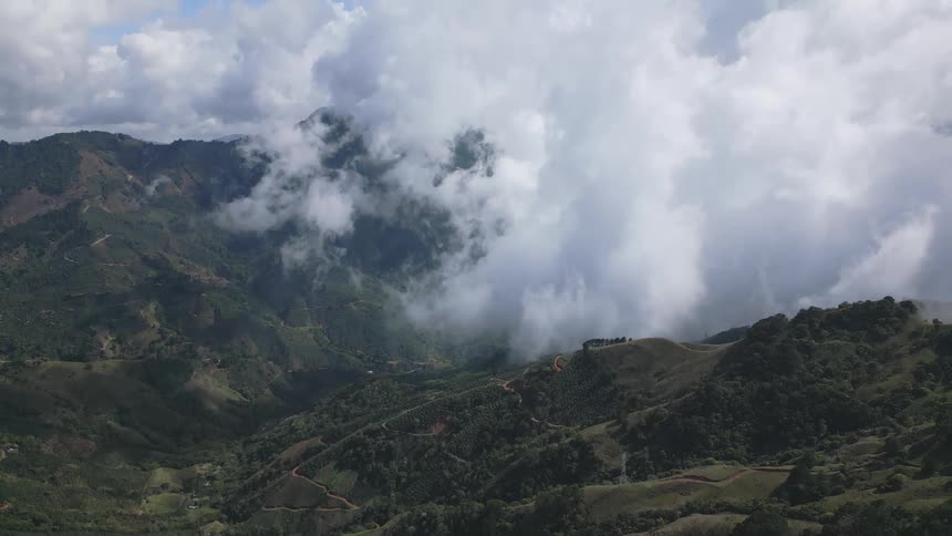 Low clouds spill over green mountain ridges revealing winding roads and valleys under a patchy blue sky