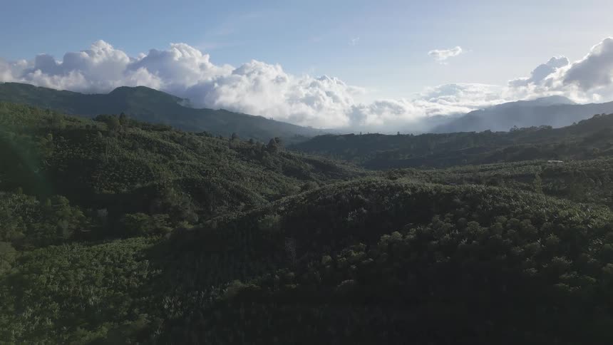 An aerial video slowly reveals a vast lush green jungle mountain range and valley with large white clouds hanging low and moving through the distant peaks