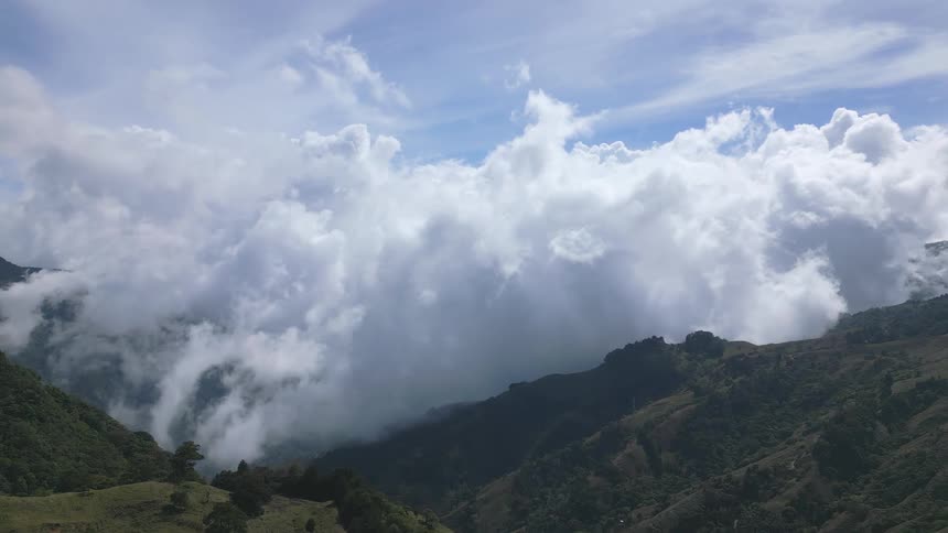 Timelapse footage shows white voluminous clouds settling over steep green mountain ridges under a bright blue sky