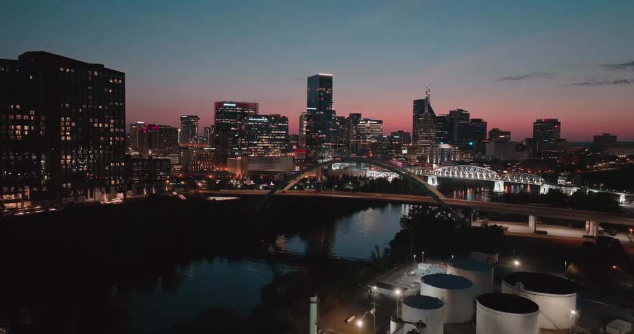 Aerial view of a city skyline at night with industrial tanks beside a river