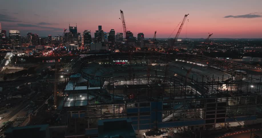 Aerial view of a stadium under construction with cranes and city skyline at dusk