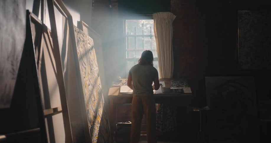 Shafts of sunlight stream through a small window into a dark dusty artists studio with exposed brick walls illuminating a desk with papers and large canvases leaning against the wall as a person walks into the shadowy foreground