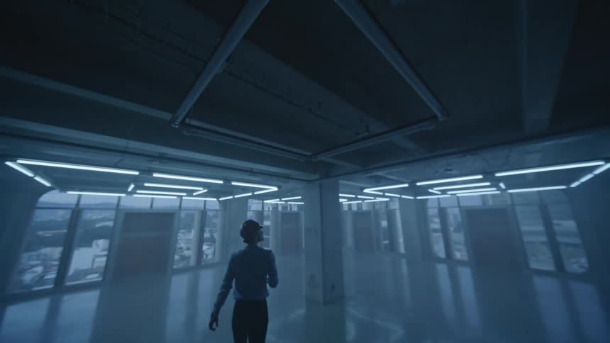 A dark wide shot of an empty large industrialstyle room with tall windows shows a silhouetted person wearing a construction hard hat standing in the center the room suddenly brightens as a grid of overhead fluorescent lights switches on il