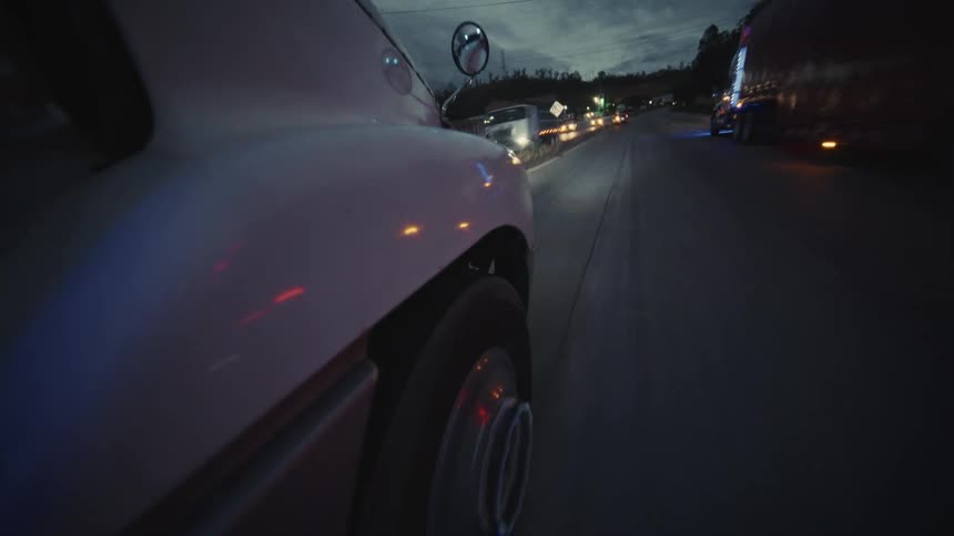 A unique lowangle shot is mounted near the rear wheel of a white vehicle driving along a highway at dusk or night the camera captures the motion of the wheel and the lights of oncoming and passing traffic on the dark road ahead