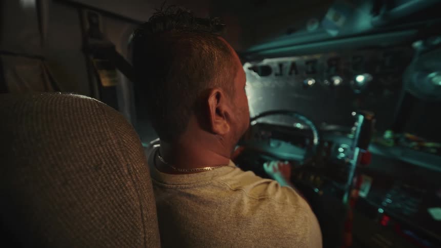 A man is sitting at the drivers seat in a semi at night lit by dashboard glow appearing focused and calm