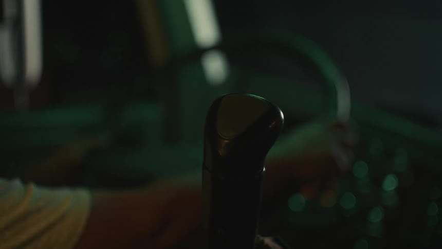 A dark moody closeup shot focuses on a cars gear shift and a drivers hand the hand approaches and then shifts the lever with the interior dimly lit by subtle green and yellow dashboard lights