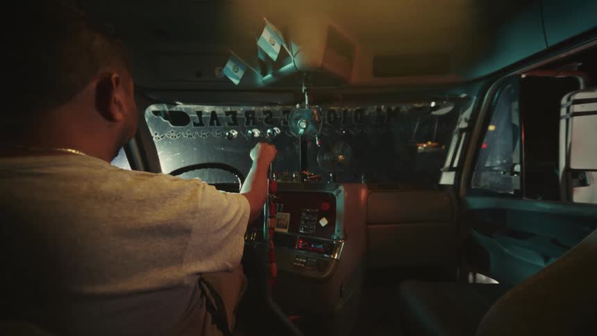 An interior view from the passenger seat shows a man driving a truck at night holding the steering wheel while small guatemalan flags hang above the windshield illuminated by dark blue cabin lights and the faint glow from the dashboard