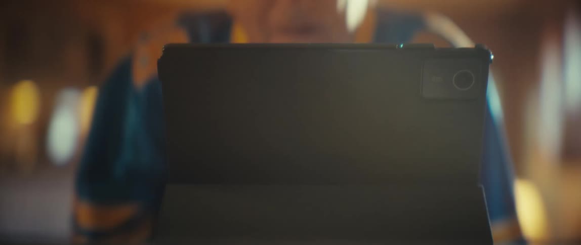 A tight closeup focuses on the back of a tablet sitting in a case on a table in the softfocus background a person wearing a blue and yellow sports jersey is looking down at the screen with a bright lens flare briefly appearing