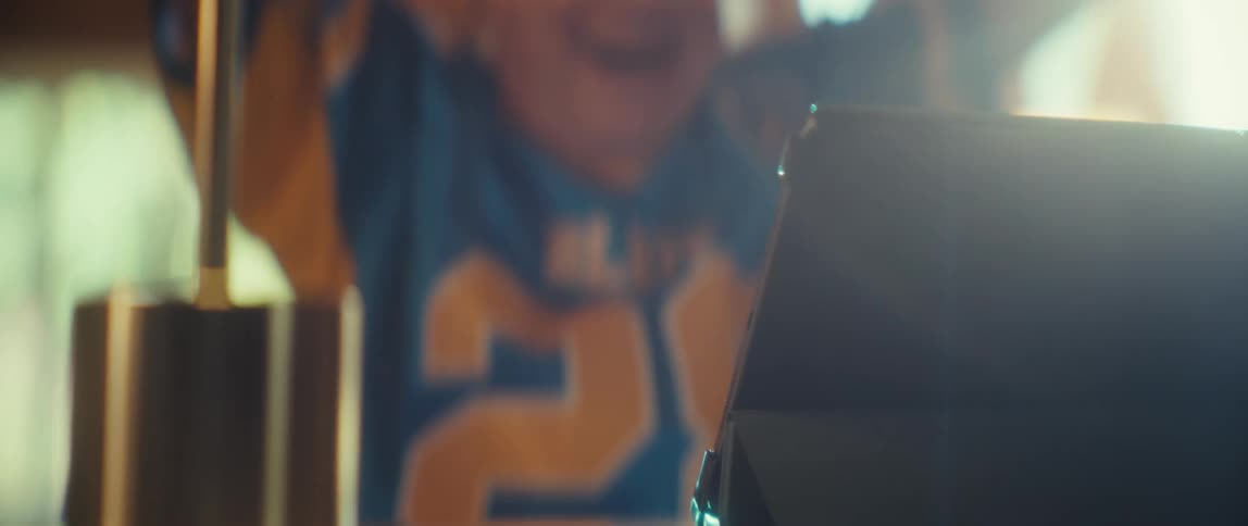 An elderly man wearing a sports jersey sits in front of a tablet clapping and cheering in a sun lit room