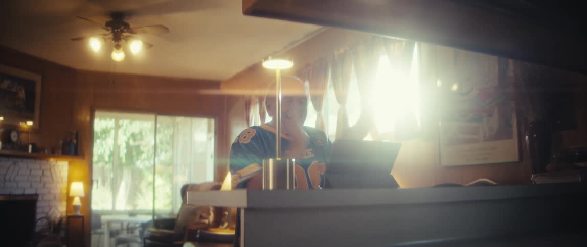 An older male wearing a sports jersey shouts and gestures excitedly while sitting at a counter and looking at a portable digital screen inside a dimly lit room with bright window light