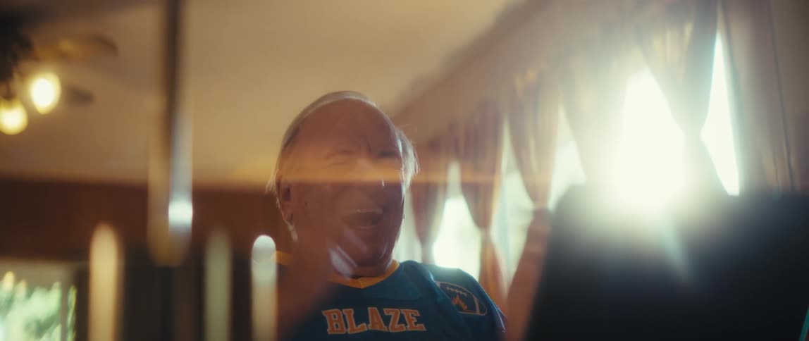 A slightly obstructed bright shot focuses on an older man wearing a blue and yellow sports jersey he is looking down at a screen unseen with a huge openmouthed smile and a cheerful expression suggesting delight and excitement